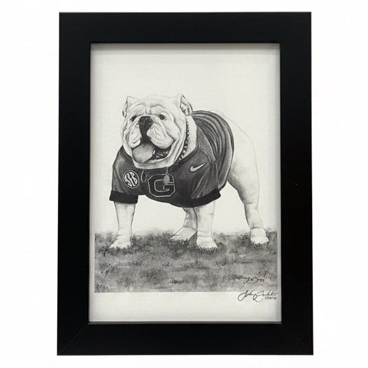 A framed black and white pencil drawing of an English bulldog wearing a football jersey, set in a black frame and leaning against a white wall on a patterned rug.