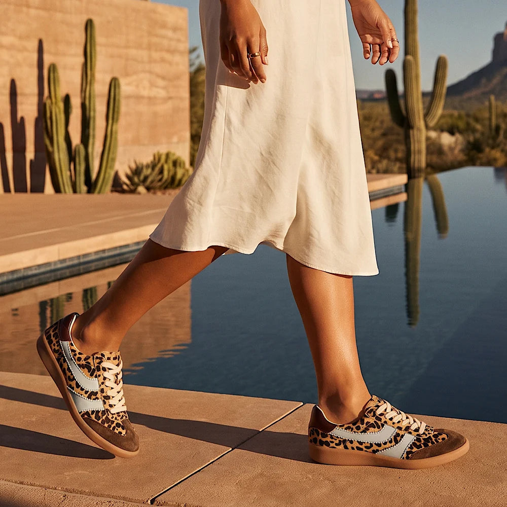 A person walking beside a pool wearing leopard-print sneakers with a light blue side stripe, in a desert outdoor setting with cacti and a stone wall.