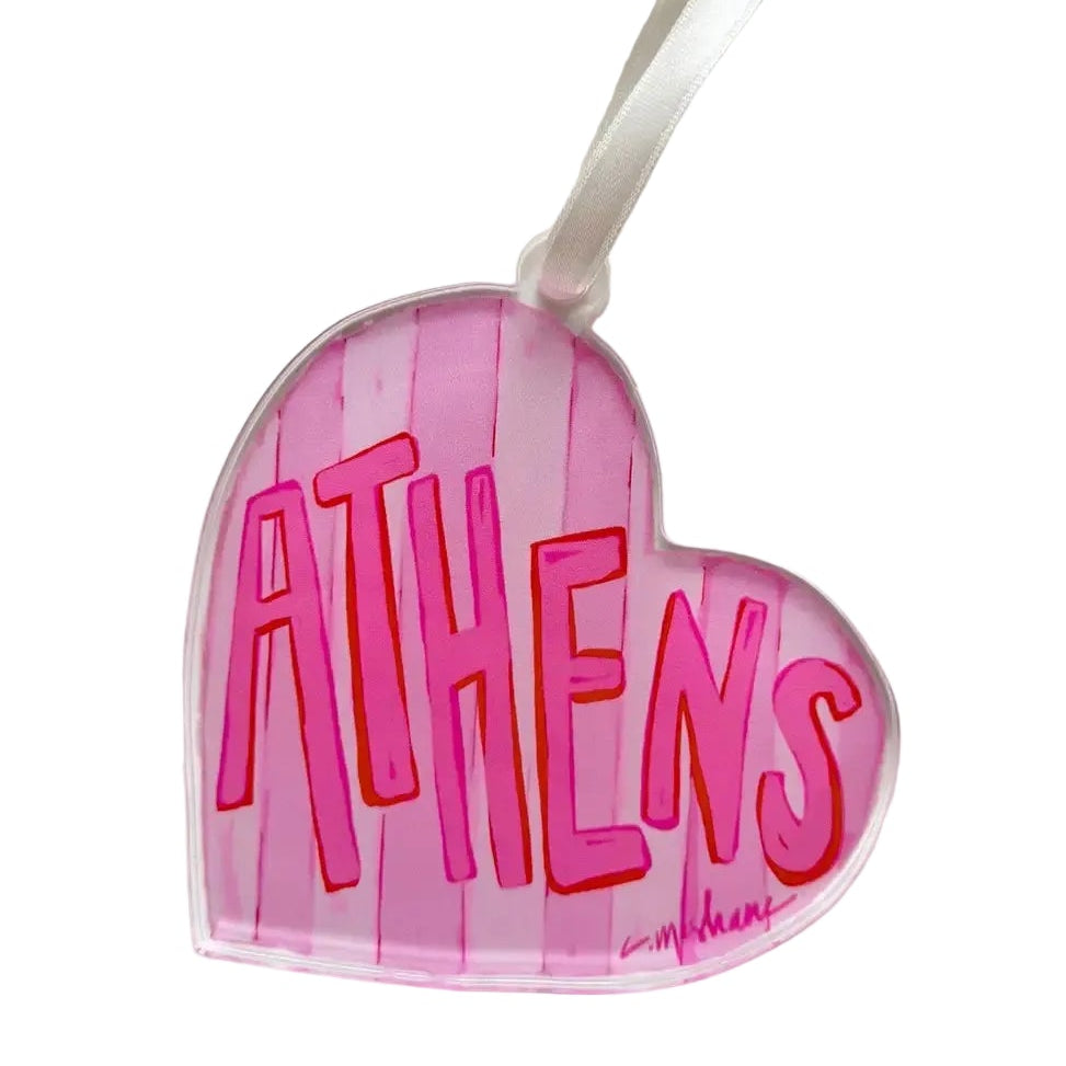 A heart-shaped clear acrylic ornament featuring the word 'ATHENS' in bold pink letters with red outlines, set against a pink striped background. A white hanging ribbon is attached at the top, and a small artist signature is visible in the lower right corner.
