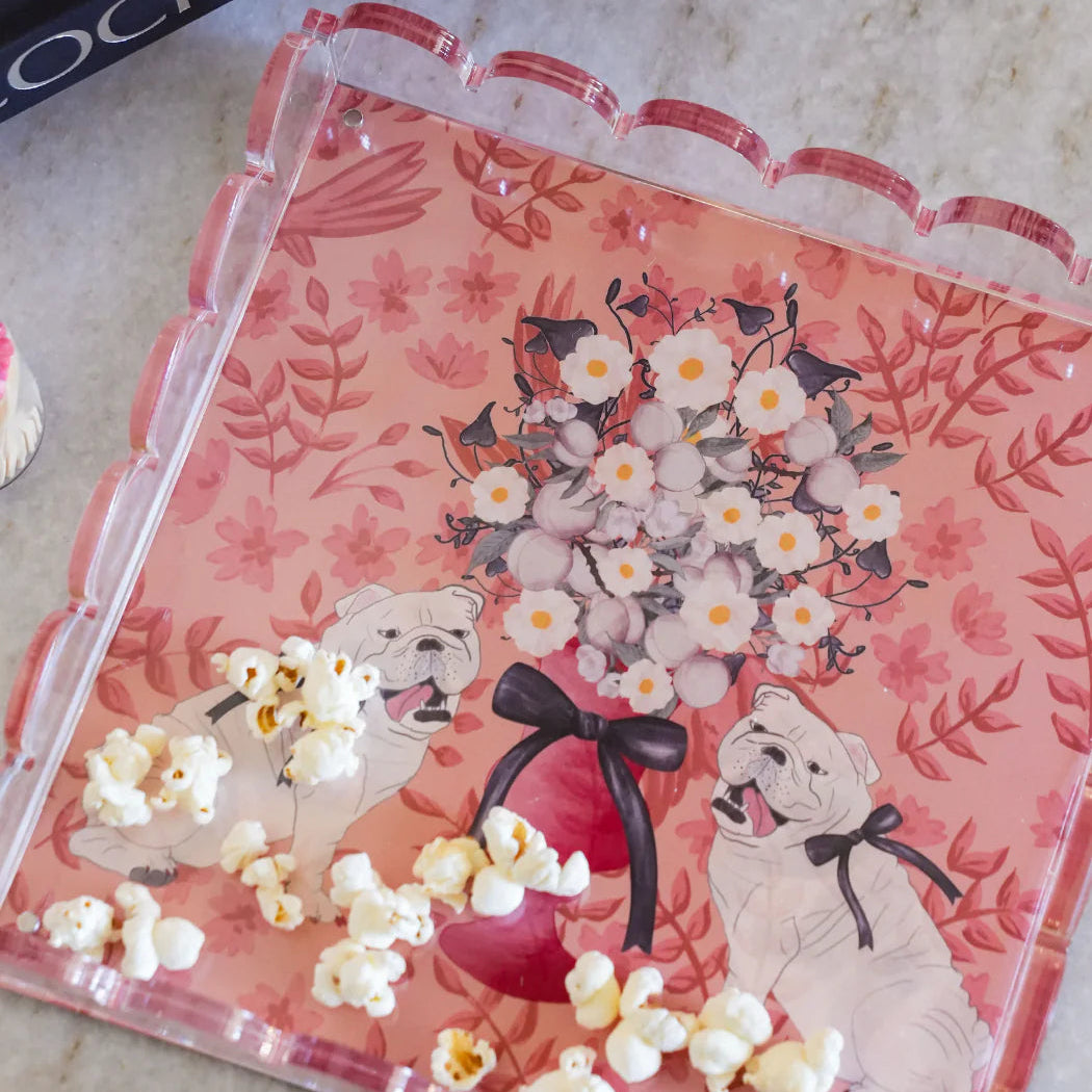 A pink scalloped acrylic serving tray with a floral and dog illustration, filled with popcorn, placed on a light marble surface with a bowl and books nearby.