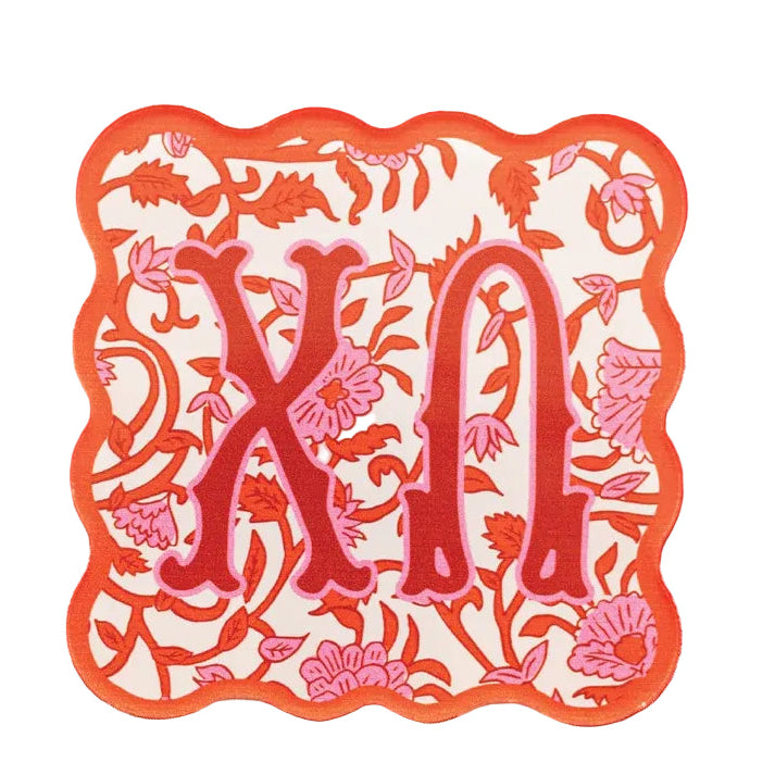 A scalloped monogram patch featuring the Greek letters Xi and Om in red, surrounded by pink floral and leaf patterns on a cream background, with a bright orange border. The patch is displayed flat on a red and white checkered fabric background.