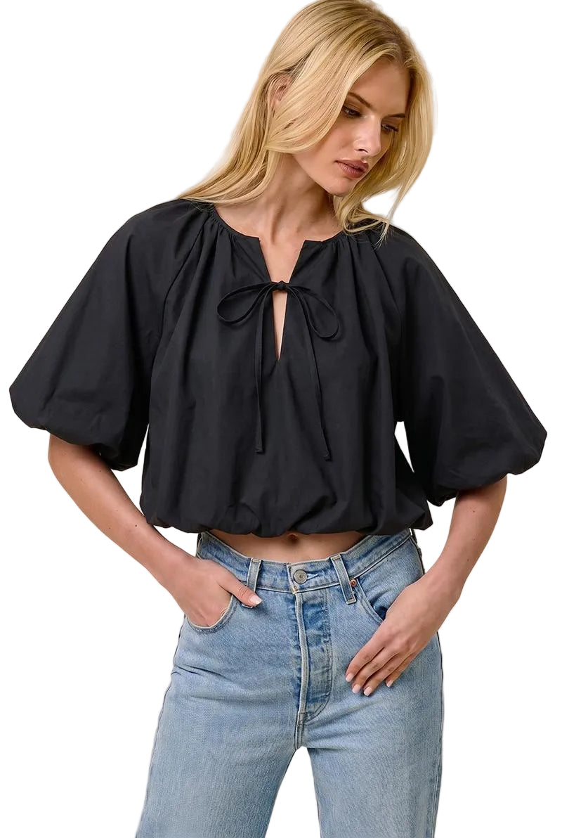 A woman wearing a black puff-sleeve blouse with a tie neckline and light-wash blue jeans, standing against a plain beige studio background.