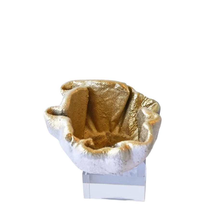 A small sculptural bowl or cup with a textured, organic shape in metallic gold and silver tones, displayed on a clear rectangular pedestal against a plain white background.