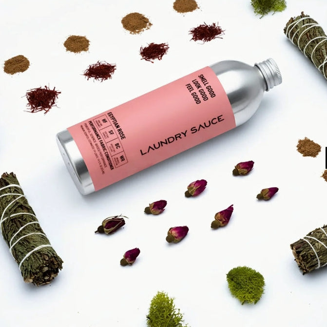 A silver bottle of laundry sauce with a pink label lies on a white surface, surrounded by dried botanicals, rose petals, and small piles of herbs. The label reads 'LAUNDRY SAUCE' and includes product details.