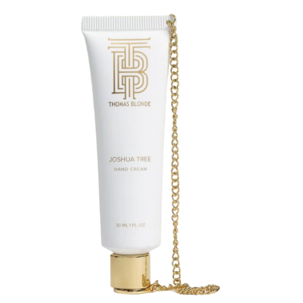A white squeeze tube of Tracee Slore Joshua Tree Hand Cream with a gold cap, featuring a gold chain draped over the side and a gold monogram logo on the front.