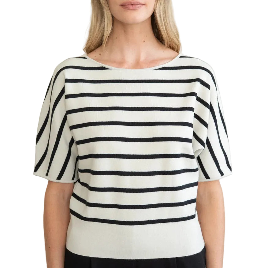 A woman modeling a short-sleeve knit top with horizontal black and off-white stripes, paired with black trousers, against a plain light beige wall.