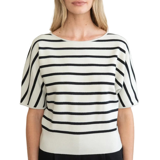 A woman modeling a short-sleeve knit top with horizontal black and off-white stripes, paired with black trousers, against a plain light beige wall.
