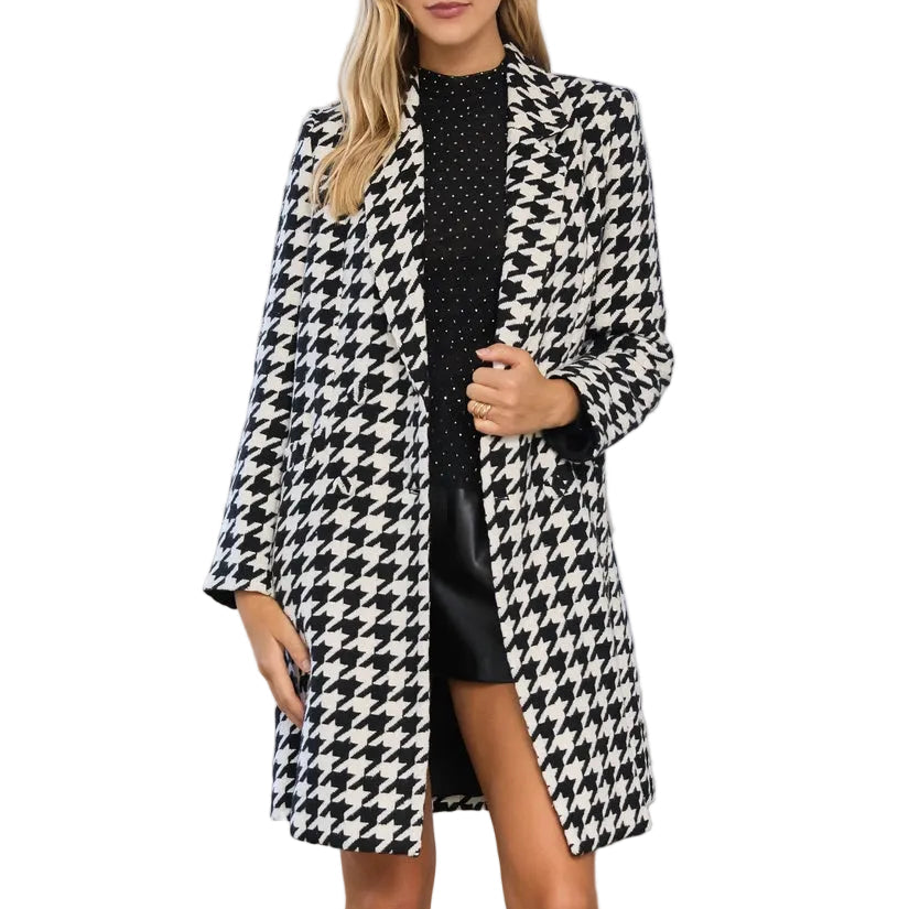 A woman models a black and white houndstooth blazer over a black mini dress, standing in a bright, white-walled room with classical columns and arches.