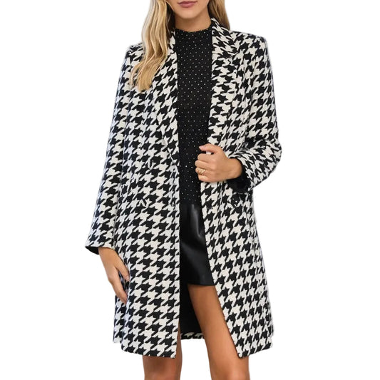 A woman models a black and white houndstooth blazer over a black mini dress, standing in a bright, white-walled room with classical columns and arches.