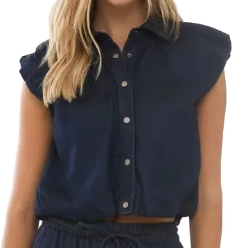 A woman modeling a navy blue sleeveless collared top and matching wide-leg trousers with a thin brown belt, standing in a bright, sunlit room with white walls and an arched window.