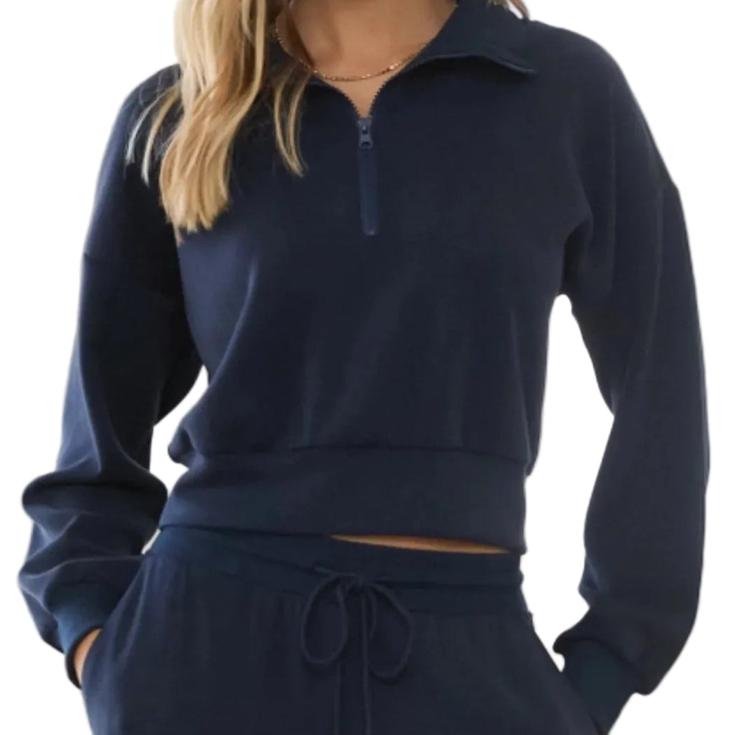 A woman wearing a matching navy blue loungewear set consisting of a cropped quarter-zip sweatshirt and drawstring sweatpants, shown from the neck down in a brightly lit indoor setting.