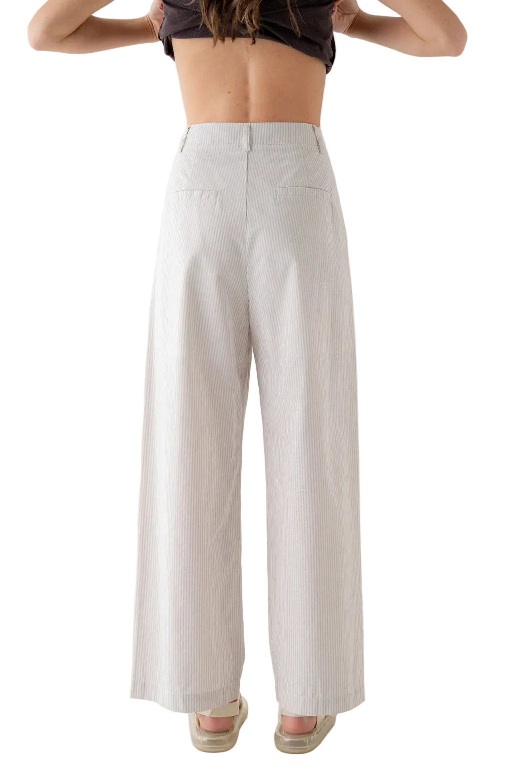 A model shown from the back wearing high-waisted, wide-leg trousers in a light beige or off-white textured fabric, paired with a dark crop top and beige sandals, against a plain white studio background.
