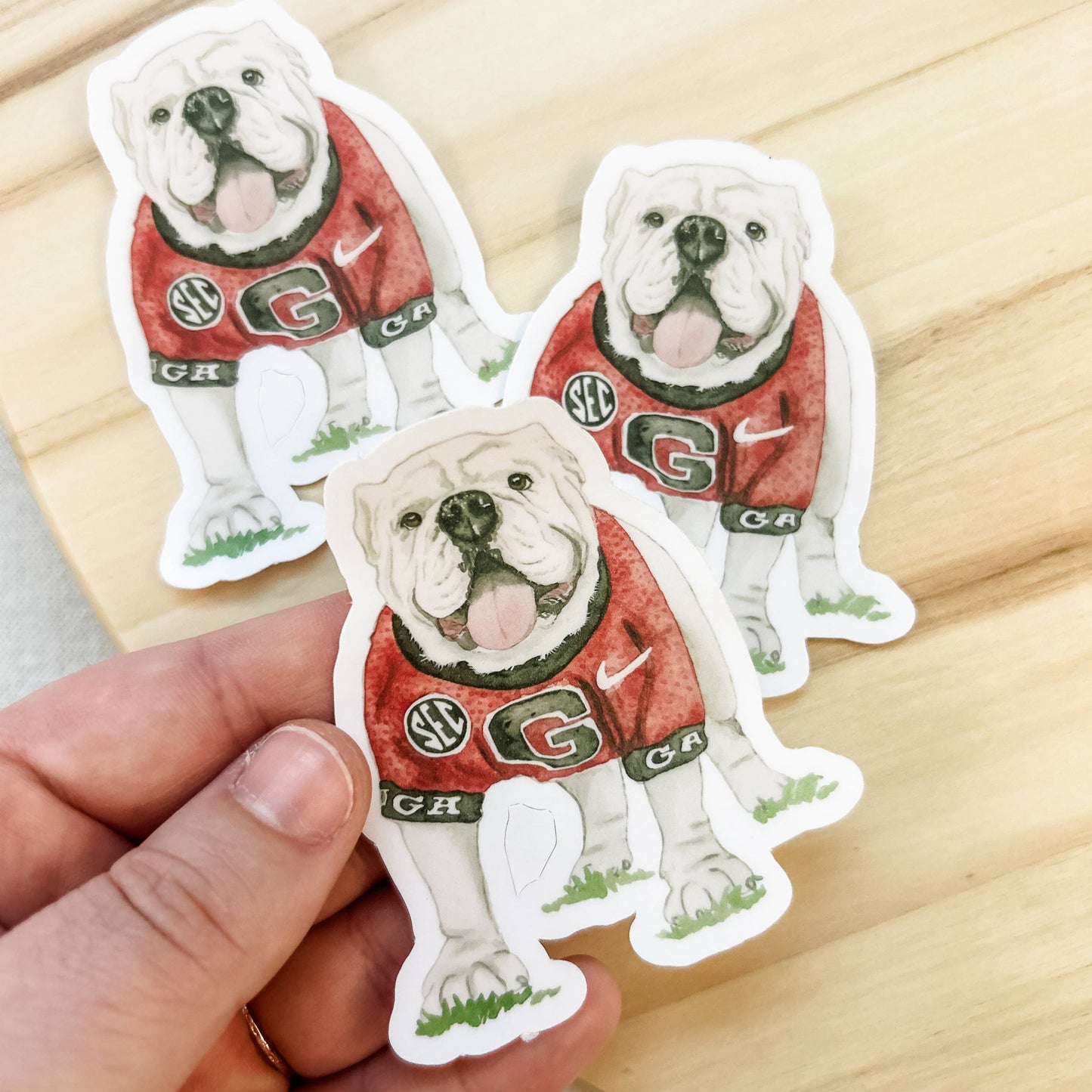 Three bulldog stickers in red and green uniforms held by a hand on a wooden surface.