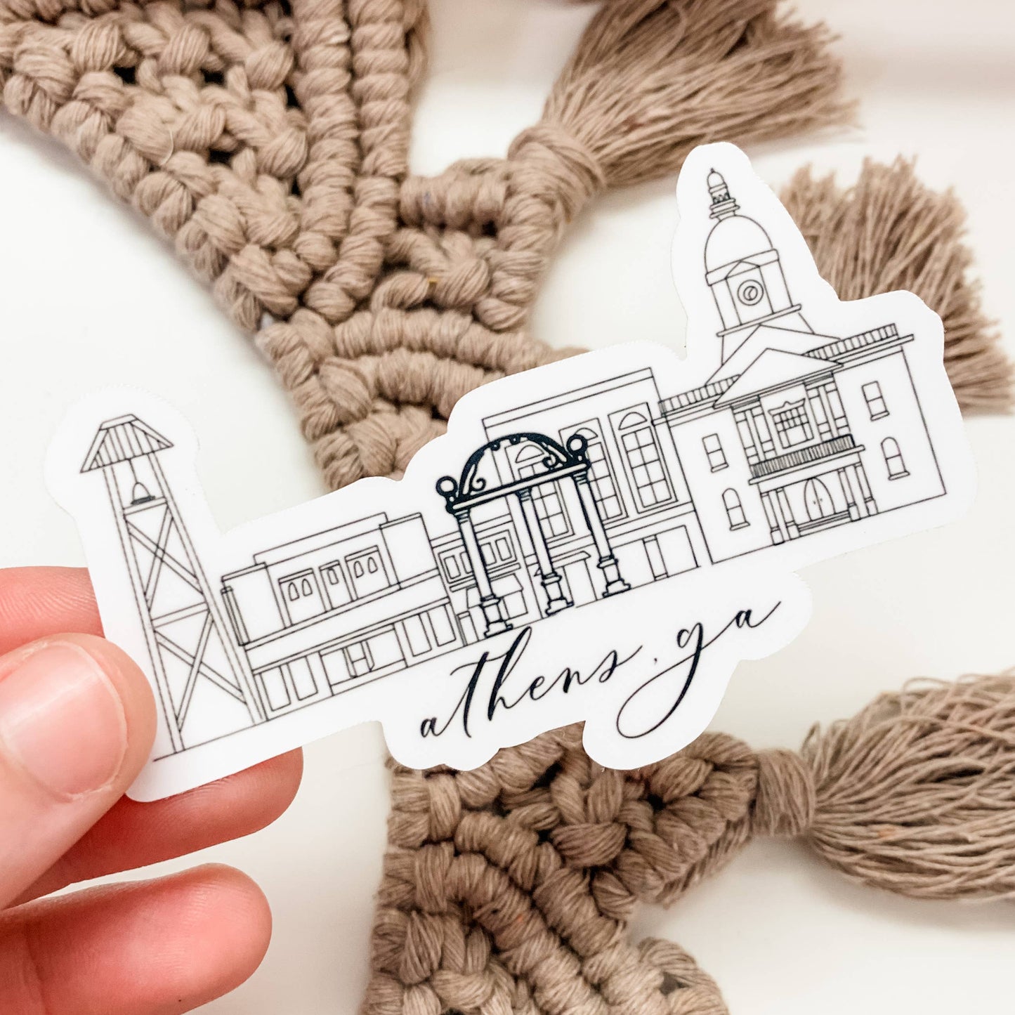 Hand holding a sticker with architectural illustrations and 'athens ga' text against a macramé background.