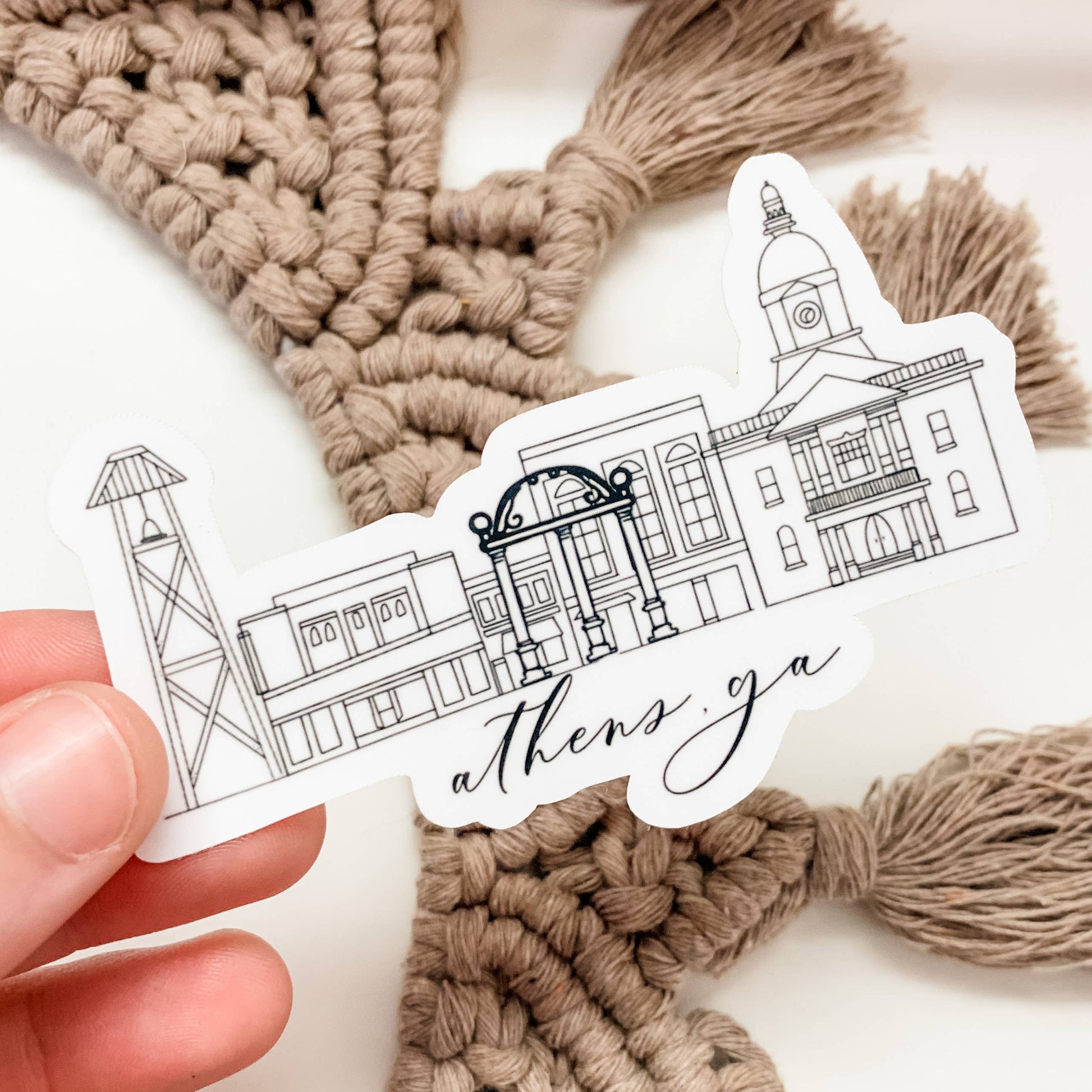 Hand holding a sticker with architectural illustrations and 'athens ga' text against a macramé background.
