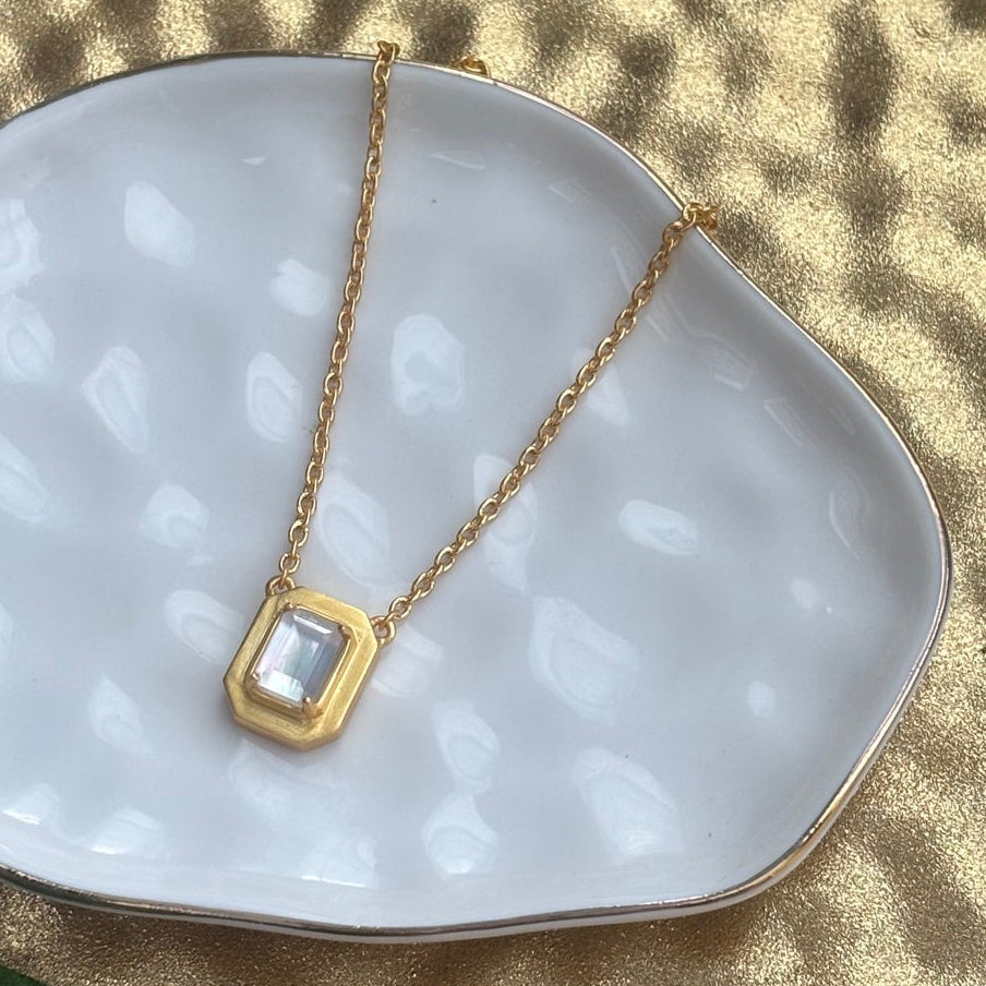 A delicate gold necklace with a small oval Mother of Pearl pendant resting on a white ceramic dish, set against a textured gold background with green leaves in the corner.