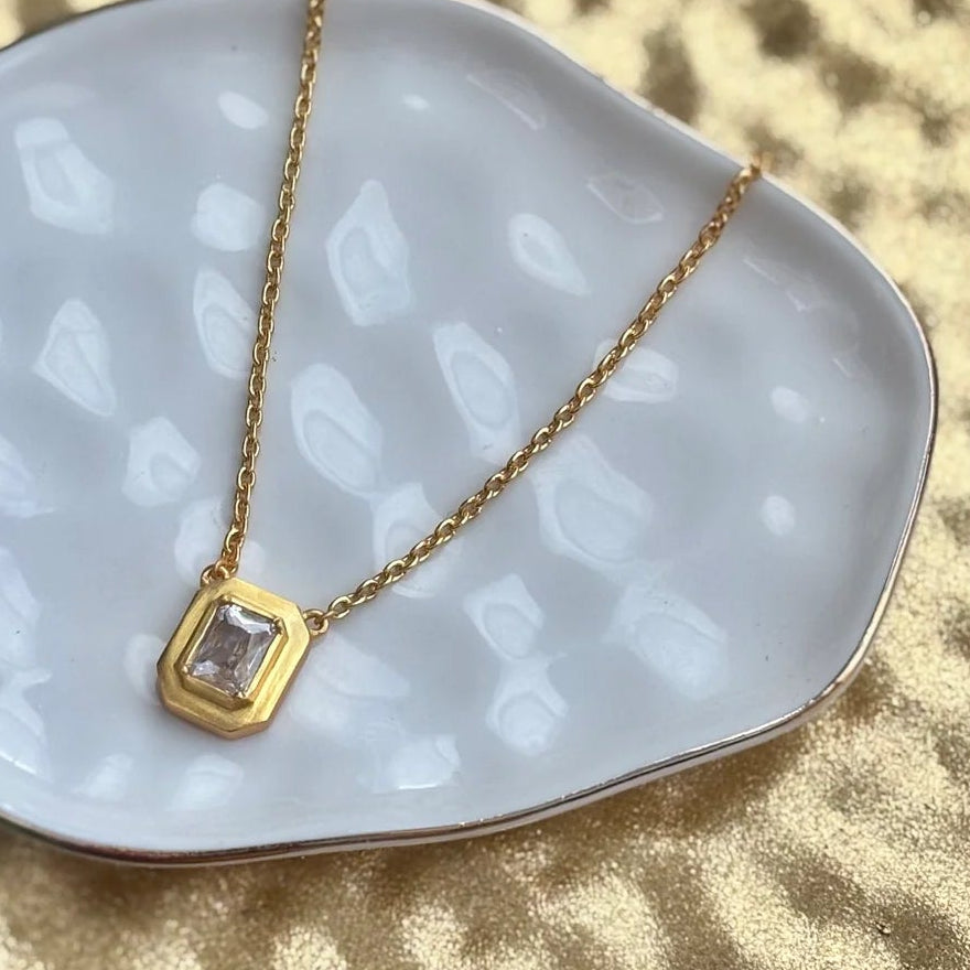 A delicate gold necklace with a small cz pendant resting on a white ceramic dish, styled on a textured gold surface with green leaves.