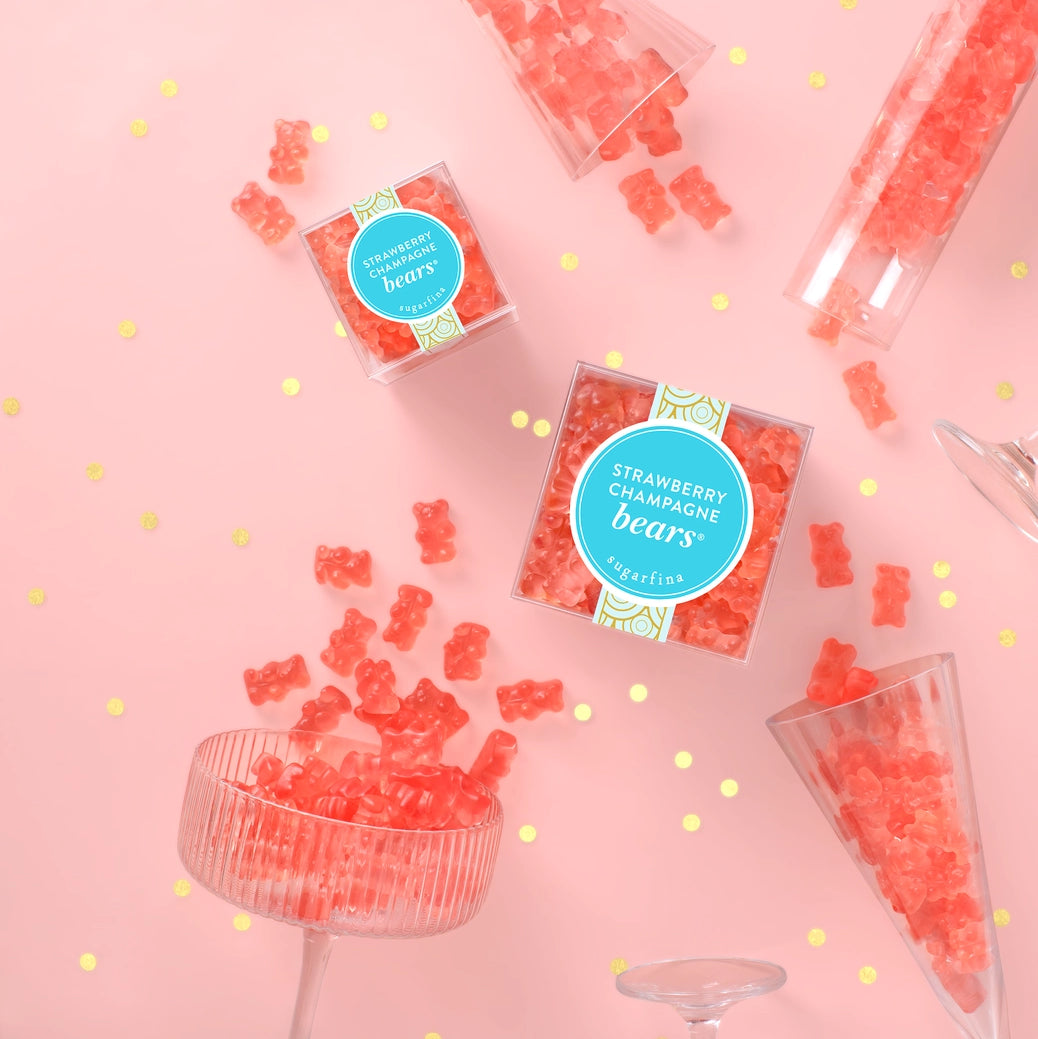 Pink packaged gummy candy bags and loose strawberry candy bears arranged on a pink background with confetti dots and glassware.