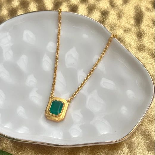 A gold chain necklace with a rectangular green gemstone pendant, displayed on a white textured dish over a gold fabric background with green leaves.