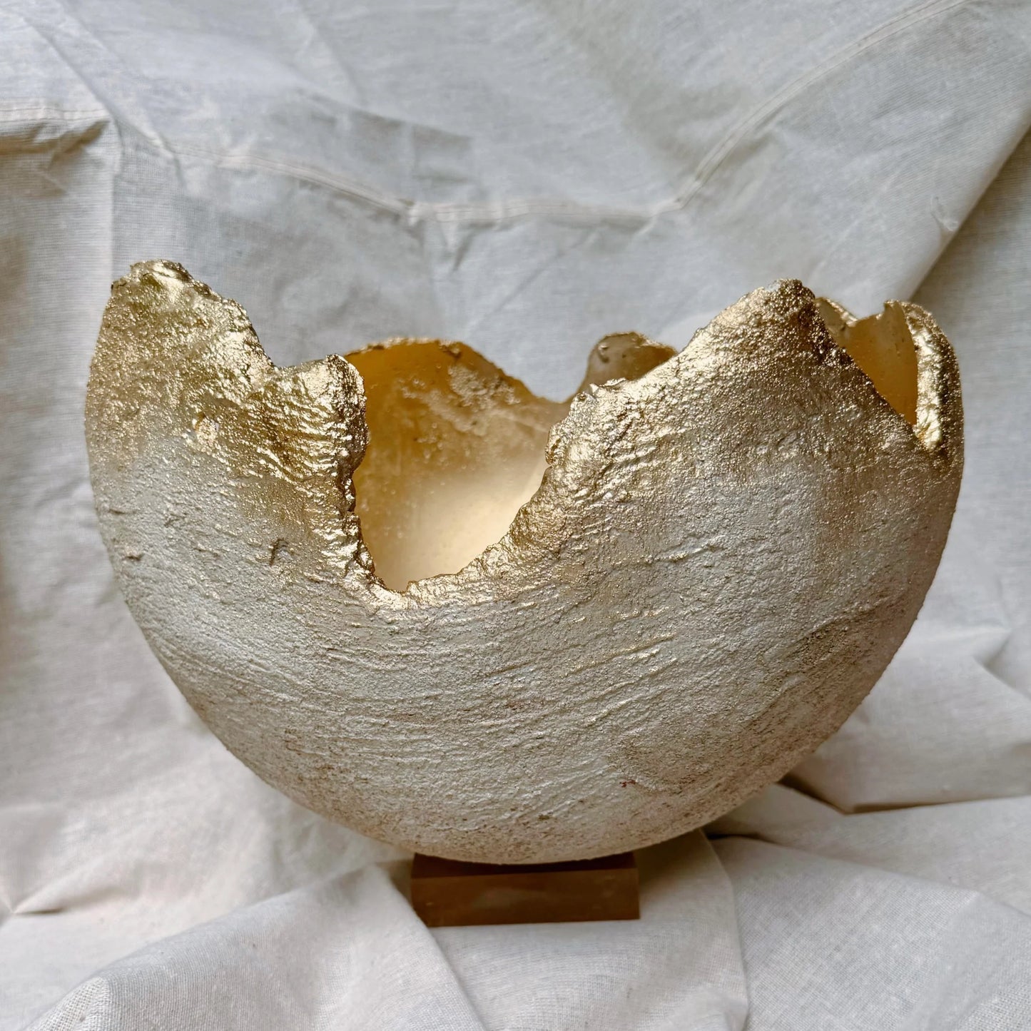 A handmade ceramic candle holder with a textured, metallic gold leaf finish, shown on a small wooden stand against a draped white linen background.
