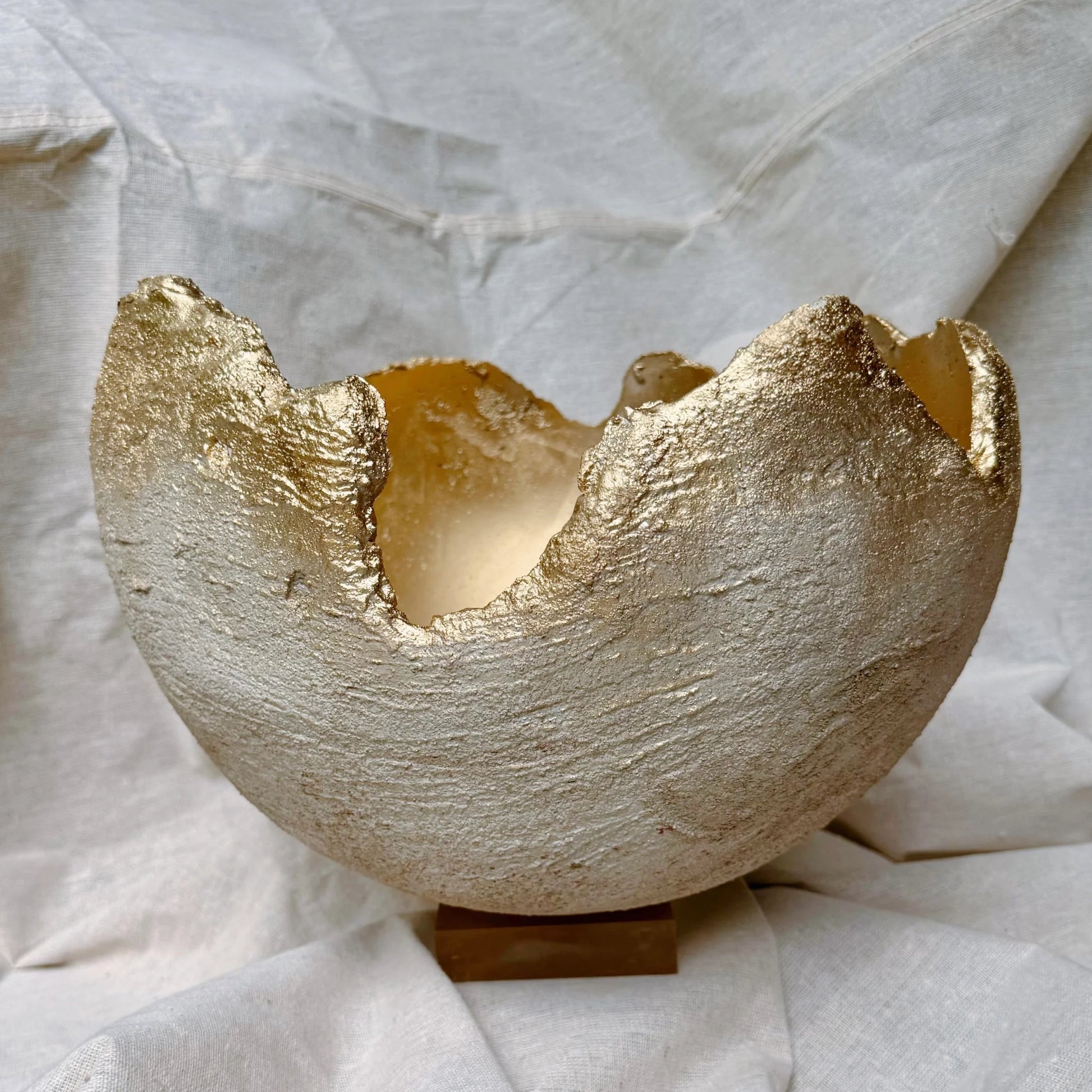 A handmade ceramic candle holder with a textured, metallic gold leaf finish, shown on a small wooden stand against a draped white linen background.