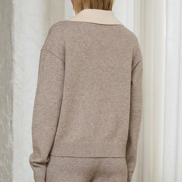 A woman stands with her back to the camera wearing a matching knit sweater and wide-leg trousers set in a light taupe-beige color, featuring a contrasting cream collar on the sweater. She is photographed in a bright, modern room with white curtains, a concrete floor, and a leather chair.