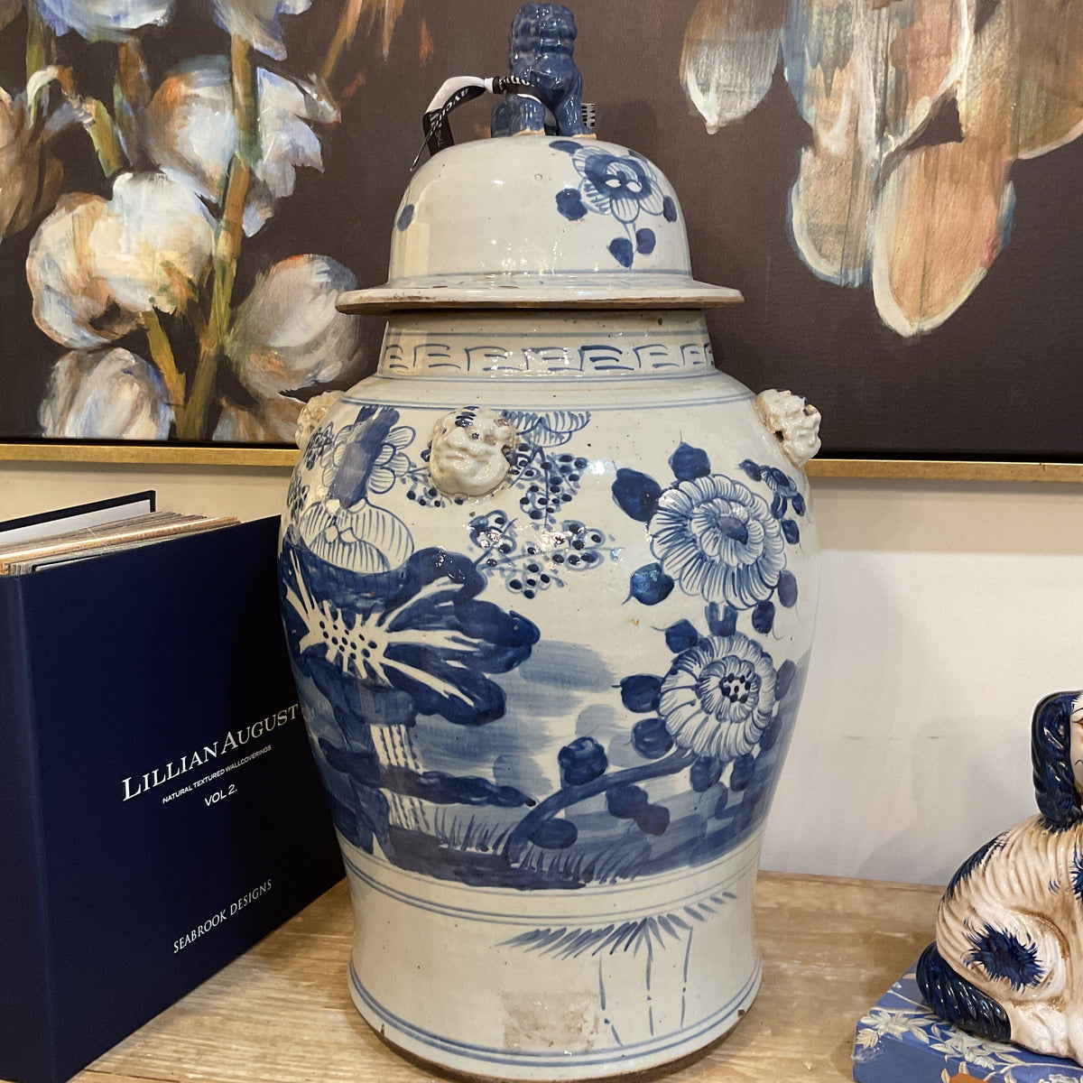 A large blue and white porcelain lidded jar with hand-painted floral and landscape motifs, displayed on a wooden shelf next to books and a painting in the background.