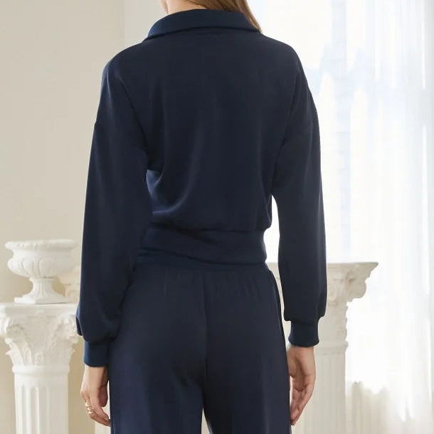 A woman viewed from behind wearing a matching navy blue long-sleeve top and wide-leg trousers set, standing in a bright, minimalist room with white pillars and an arched window.
