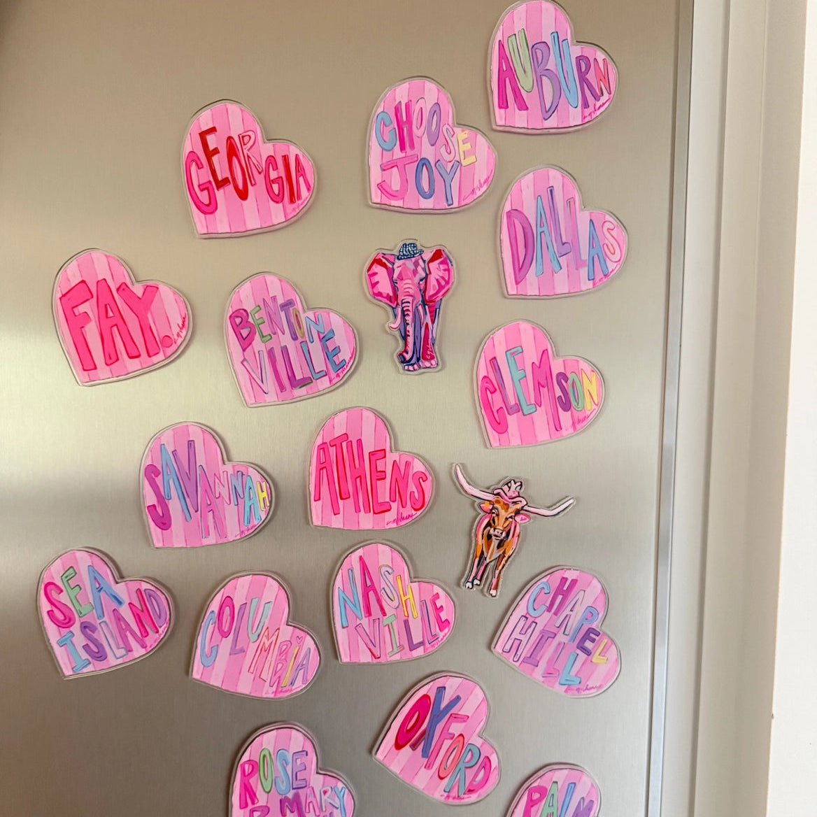A collection of hand-painted pink heart-shaped refrigerator magnets with various names and words written in colorful lettering, displayed on a metallic silver refrigerator.