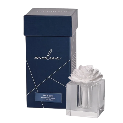 A small rectangular dark blue perfume box with white script reading 'modena' stands next to a clear glass perfume bottle topped with a white flower, on a plain white background.