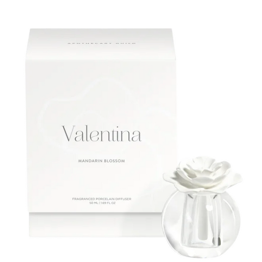 A white rectangular Valentino perfume box with the brand name printed in gray, shown next to a clear glass reed diffuser bottle with a white flower-shaped top, on a white background.