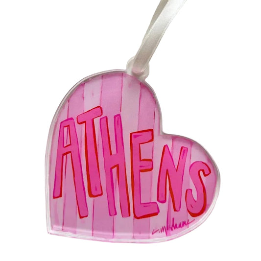 A heart-shaped clear acrylic ornament featuring the word 'ATHENS' in bold pink letters with red outlines, set against a pink striped background. A white hanging ribbon is attached at the top, and a small artist signature is visible in the lower right corner.