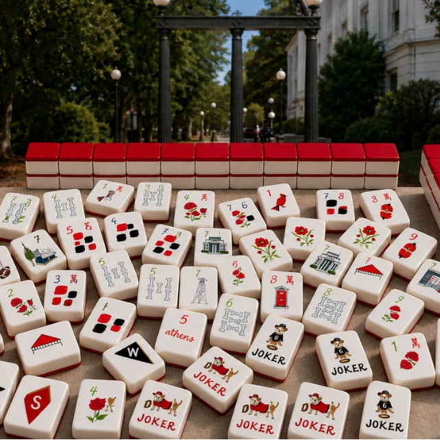 A set of Mahjong tiles arranged outdoors, with a row of red-backed tiles behind and many face-up tiles in front. The background shows a blurred park scene with trees, lampposts, and a building.