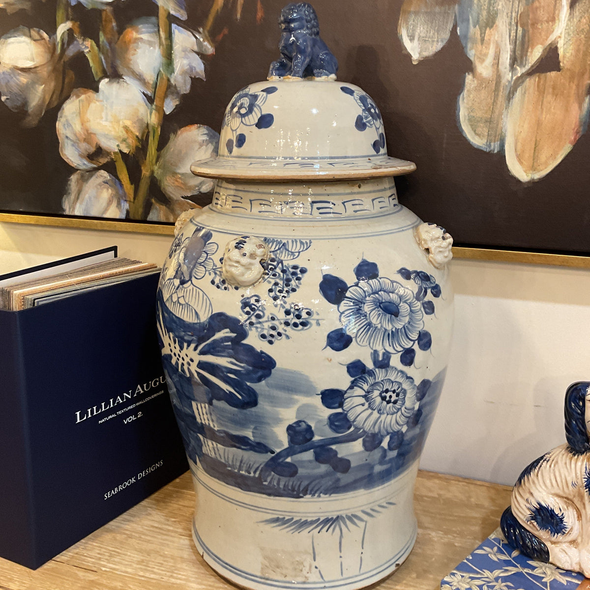 A blue and white porcelain lidded jar with floral and landscape motifs, displayed on a wooden surface next to a dark blue book and a framed painting.