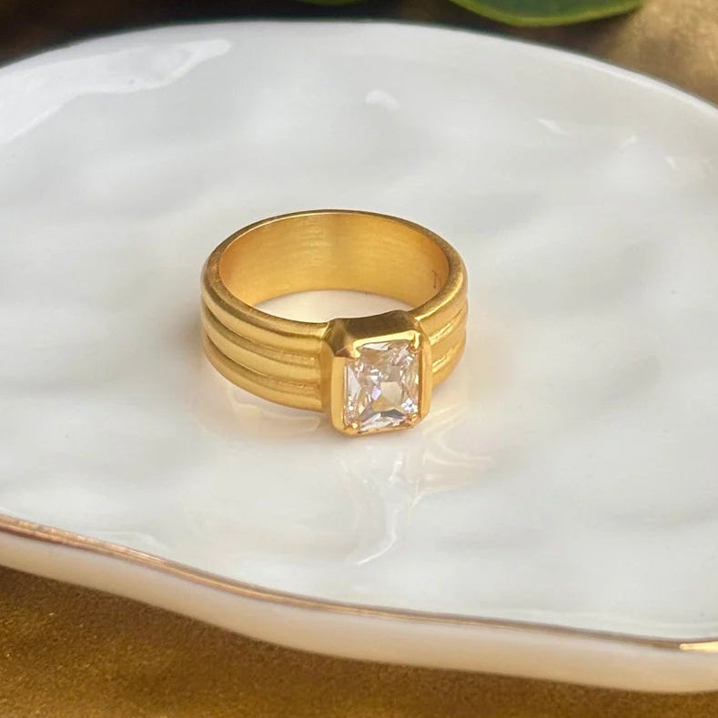 A gold ring with a small CZ gemstone set in a white clamshell jewelry display, resting on a textured gold surface with green leaves in the background.