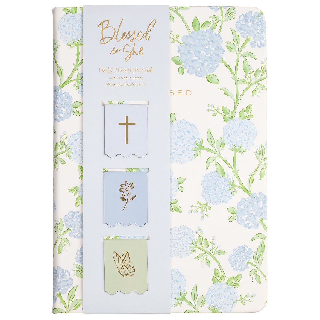 A slim journal or notebook with a floral cover design featuring light blue hydrangeas and green leaves on an off-white background. The left side has a pale blue vertical panel with gold foil text reading 'Blessed is She' and three small decorative flag-like inserts with a cross, a flower, and a peace sign.