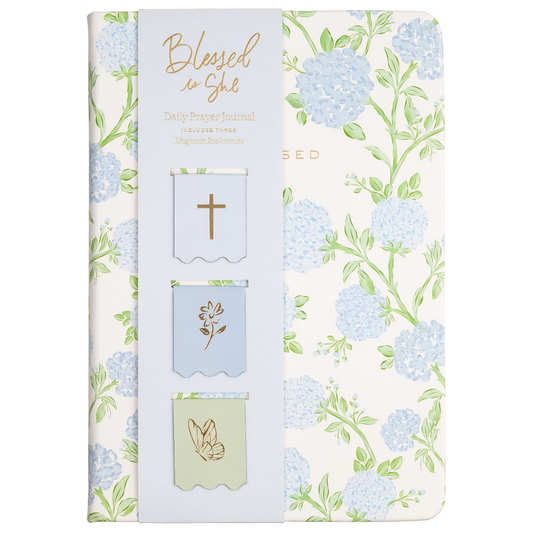A slim journal or notebook with a floral cover design featuring light blue hydrangeas and green leaves on an off-white background. The left side has a pale blue vertical panel with gold foil text reading 'Blessed is She' and three small decorative flag-like inserts with a cross, a flower, and a peace sign.