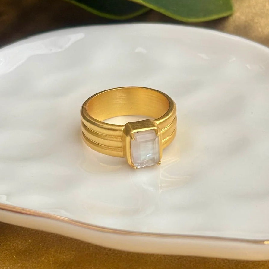 A gold ring with a rectangular Mother 0f Pearl stone sits on a white ceramic dish with gold trim, styled with green leaves on a textured gold surface.