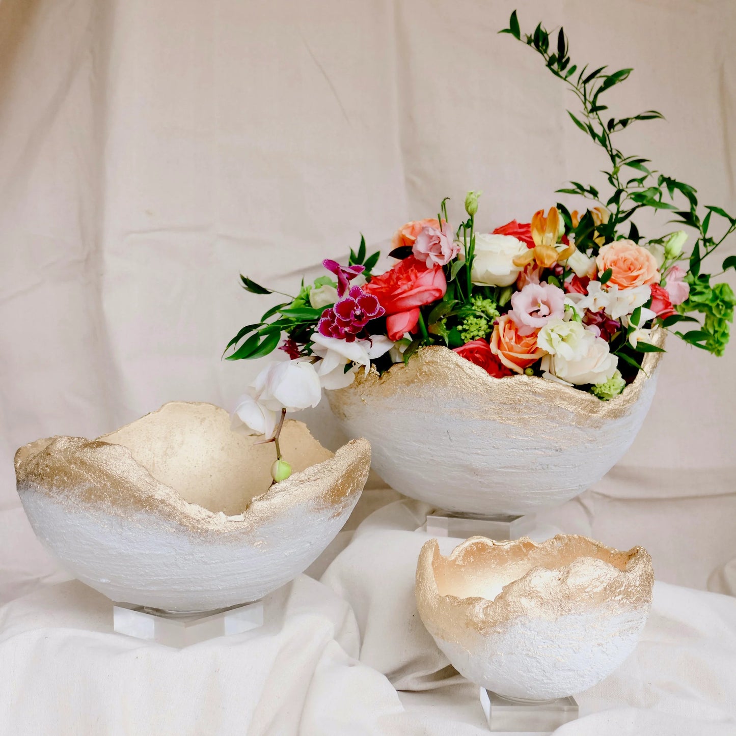 Three rustic white ceramic egg-shaped bowls with gold glittery rims, displayed on a draped white fabric surface. One bowl is empty, while the other two are filled with a colorful floral arrangement featuring red, pink, orange, and white flowers with green leaves.