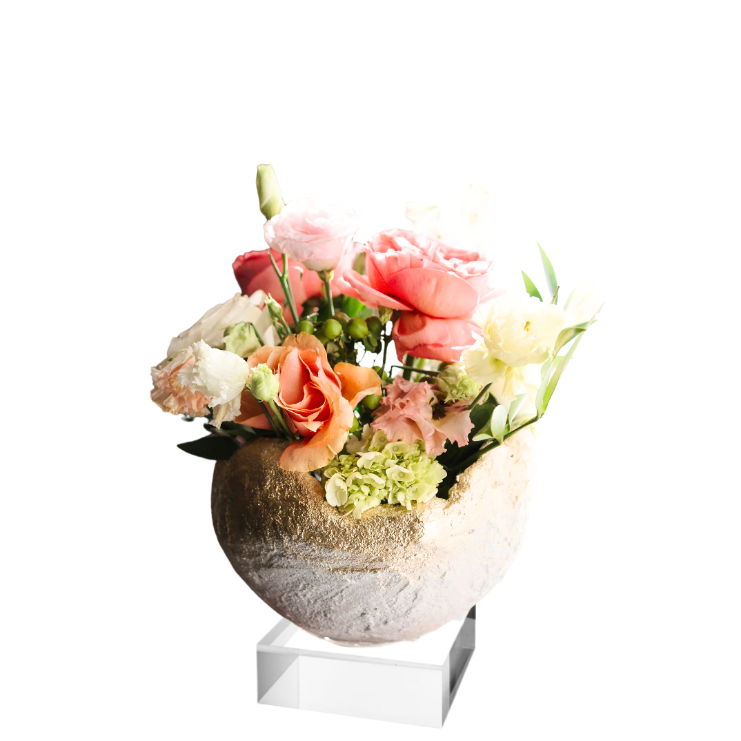 A floral arrangement in a textured round vase with a clear acrylic pedestal stand, set against a solid white background.