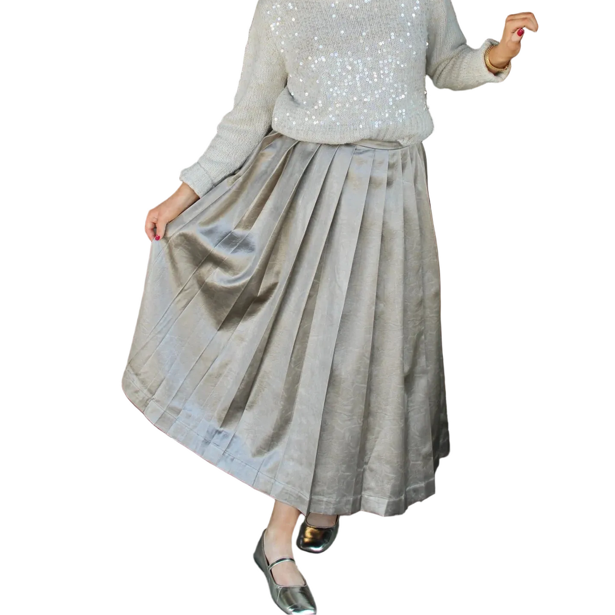 A woman wearing a sparkling light beige sequined sweater and a pleated taupe skirt stands in a storefront doorway, holding the door open with one hand while holding out the skirt to show its drape.