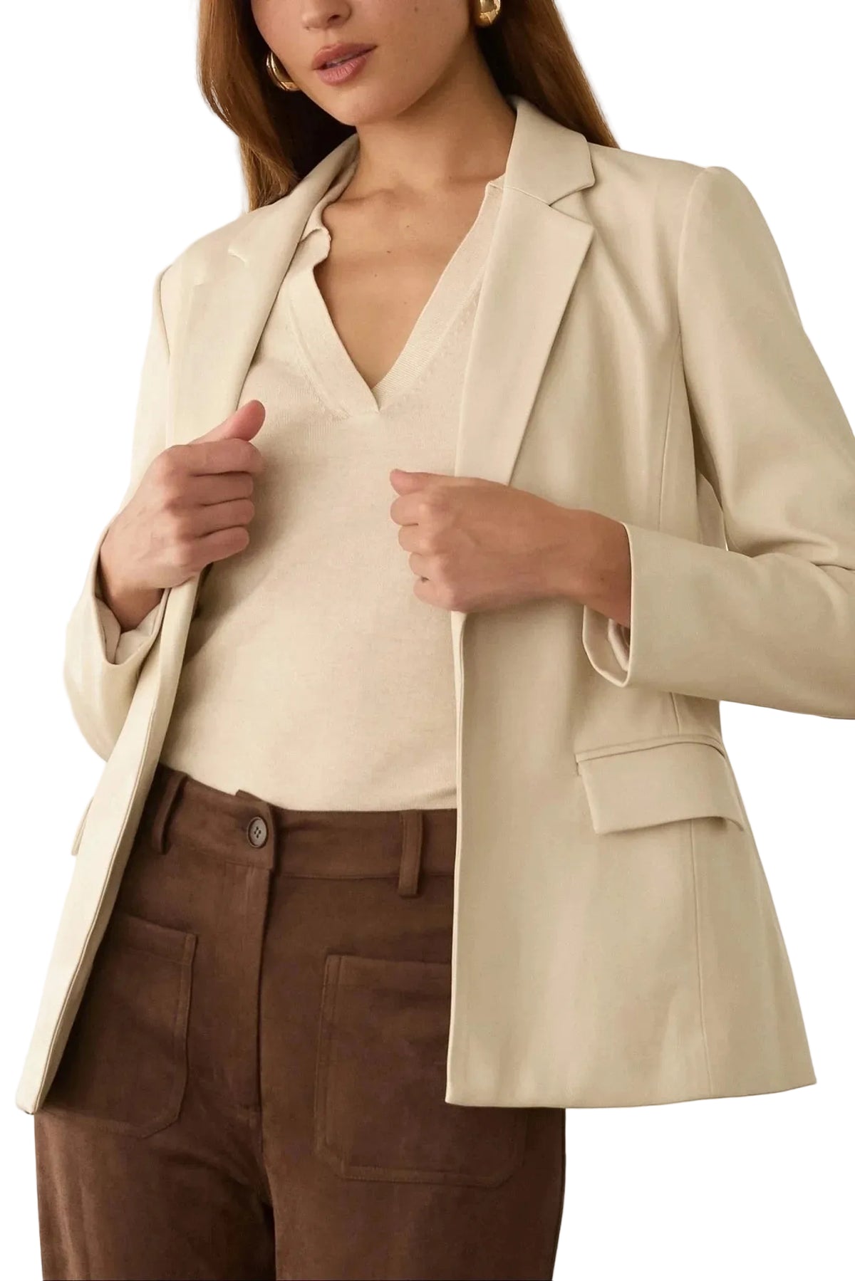 A woman wearing a light beige blazer over a matching beige V-neck top and brown pants, shown from the shoulders to the hips in an indoor setting with a light cream wall background.