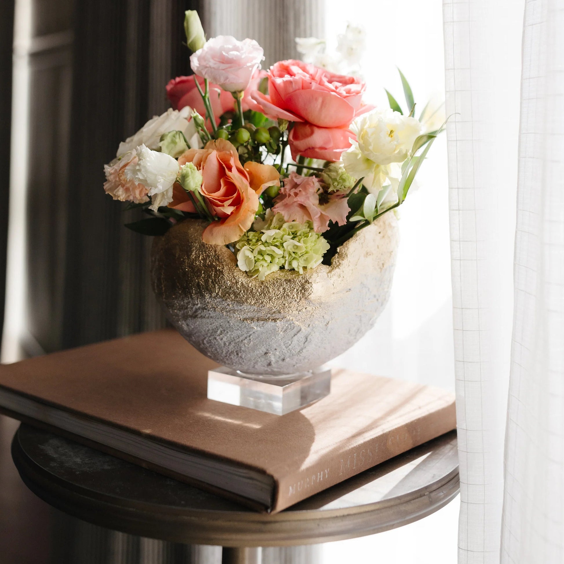 A floral arrangement in a textured round vase resting on a brown book on a small round table, set against a softly lit window with sheer white curtains and dark drapes.