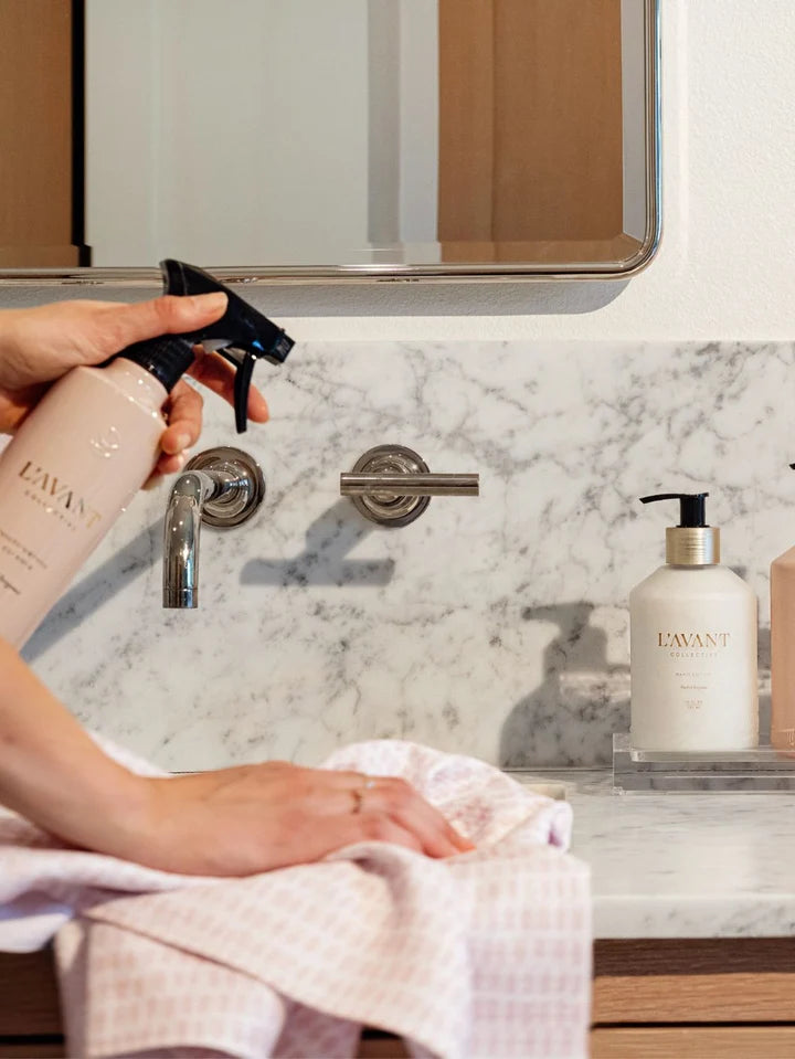 A person's hands are shown holding a spray bottle of EWAN bathroom cleaner and wiping a marble countertop with a checkered towel in a modern bathroom setting.