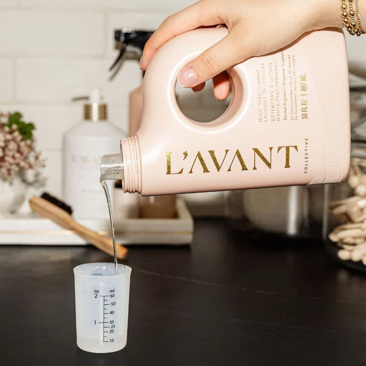 Person pouring liquid from a pink 'L'AVANT' container into a measuring cup on a kitchen counter.