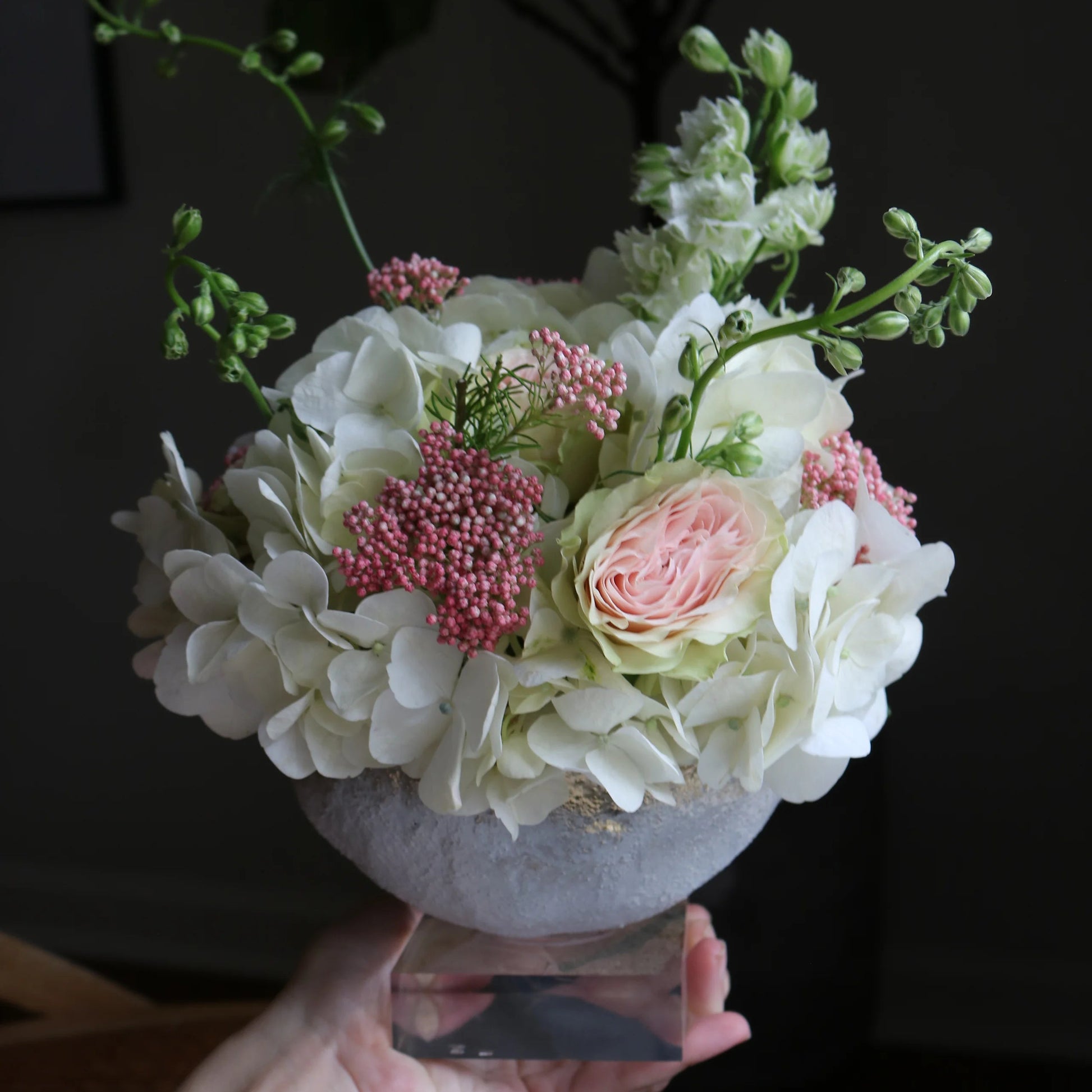 A floral arrangement in a round white bowl vase, held by a hand. The bouquet features white and light pink flowers with green stems, set against a dark, shadowed background.