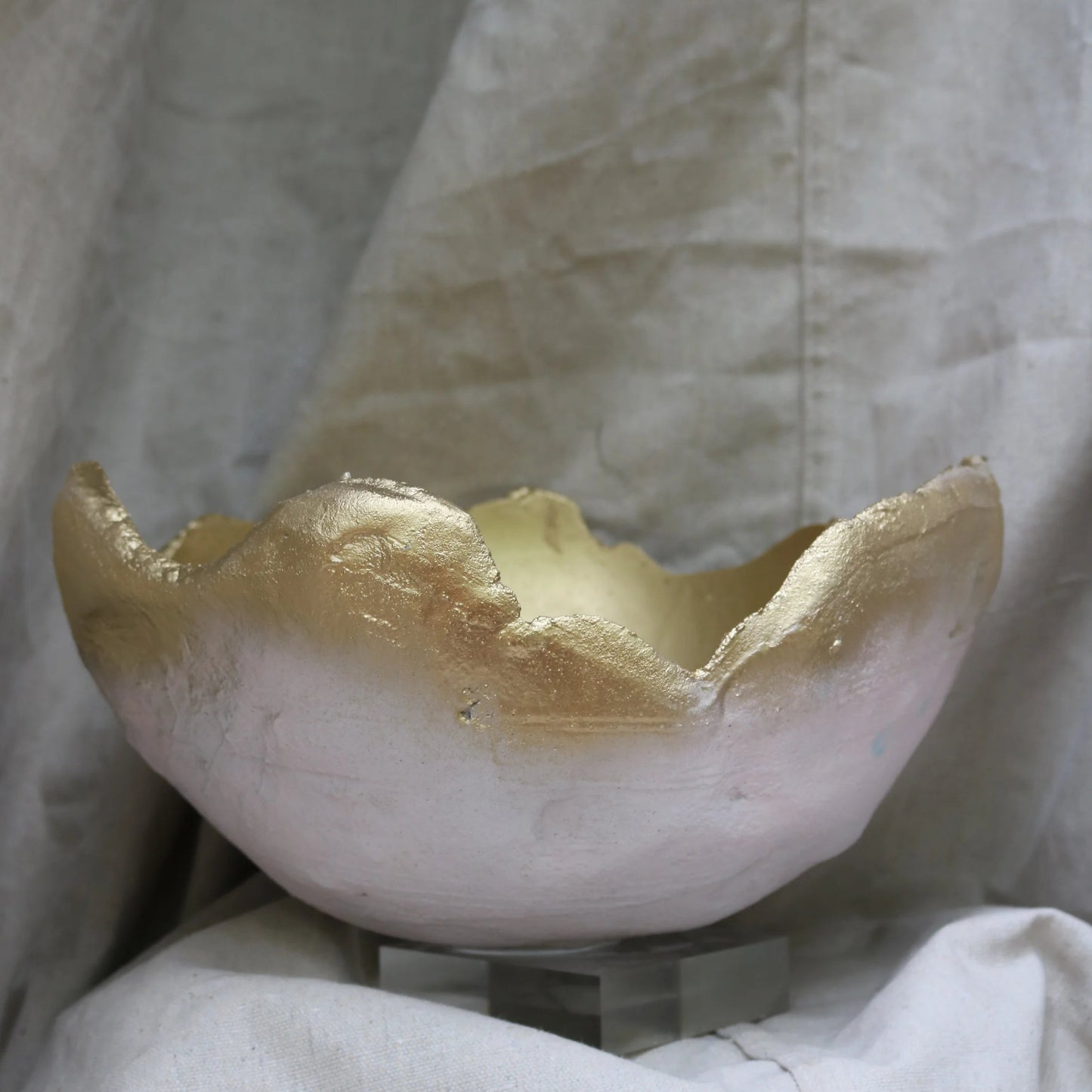 A handmade ceramic bowl with a rustic, broken-edge rim, featuring a white exterior and a metallic gold-painted interior, displayed on a small stand against a draped beige fabric backdrop.
