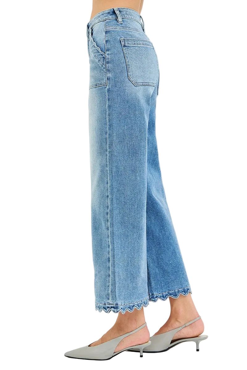 A side view of a model wearing light blue cropped wide-leg denim jeans with a scalloped hem, paired with gray slingback kitten heels, against a plain white studio background.