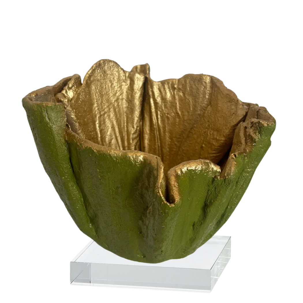 A sculptural bowl with a textured olive-green exterior and a metallic gold interior, displayed on a clear acrylic block against a white background.