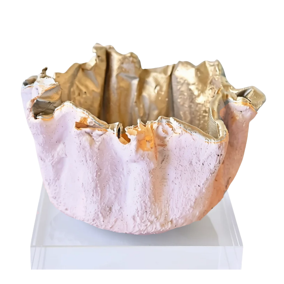 An abstract sculptural bowl with a textured pale pink exterior and metallic gold interior, displayed on a clear acrylic riser against a white background.